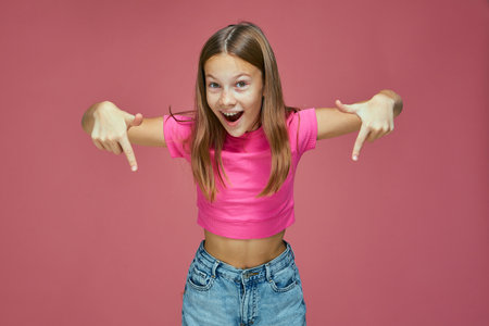 Cool offer. Surprised happy child girl point down. Excited schoolgirl blogger show link to subscribe on pink backgroundの写真素材