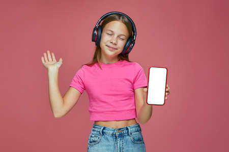 Music mobile app advertisement. Happy little girl child in headphones hold phone with white empty screen mock upの写真素材