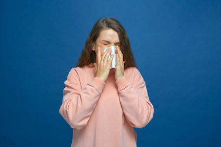 Sick young girl sneezing in tissue, blowing runny nose, suffer seasonal allergy or influenza symptoms on blue backgroundの写真素材