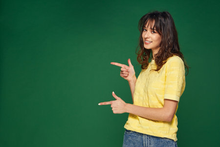 Modern beautiful young girl point to side showing on empty space on green background for sale offer,...