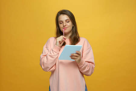 Modern pretty young girl planning schedule, make to do list in notepad on yellow studio background. Time managementの写真素材