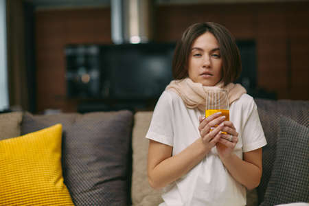 Unwell caucasian girl sit at home suffer cold or influenza, hold glass of medicines or vitamin c to relieve symptomsの写真素材