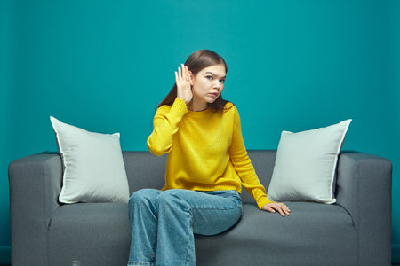 Young girl with hand near ear listen to whisper, try to understand quiet talk, having hearing problems, sitting on sofaの写真素材