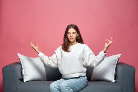 Calm young woman with eyes closed, do mudra om gesture meditating, breathing, sitting on sofa....