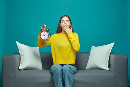 Sleepy young girl yawn, covering mouth by hand, holding alarm clock. Bedtime, jet lag, sleep deprivation conceptの写真素材