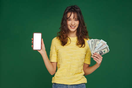 Lucky young girl hold money cash in hand and phone with blank template screen, advertise banking app on green backgroundの写真素材