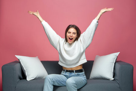 Overjoyed lucky young girl celebrating win with raised arms, screaming with euphoria, sitting on sofaの写真素材