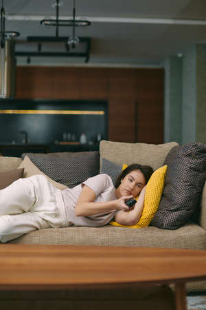 Modern young woman watching tv programs, movie or series changing channels by remote controller, lying on sofaの写真素材