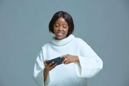 Happy cute african american young girl playing online game using modern smartphone apps toothy...