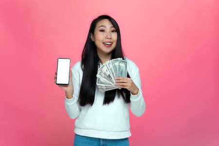 Lucky young asian girl hold money cash in hand, phone with blank mockup screen, enjoys win, jackpot on pink backgroundの写真素材
