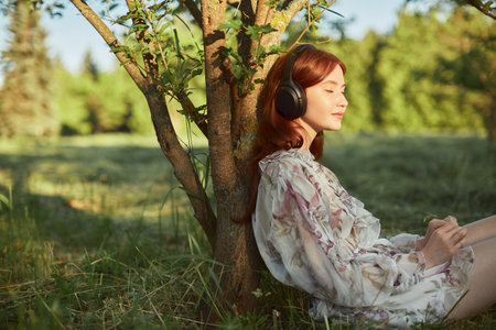 A red-haired pretty girl in headphones listens to music sitting by a tree. The rays of the setting sun fall on the face of a young woman.の写真素材
