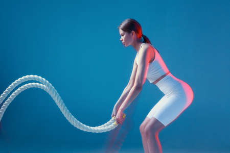 Sporty fitness girl pushing sports ropes on blue backdrop with copy space. Sports club advertising....