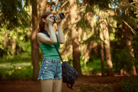 Pretty young red-haired woman in a green top and denim shorts takes pictures in the woods.の写真素材
