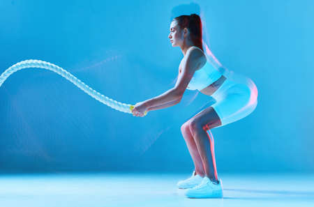 Sporty pretty tanned young woman exercises with rope. Fitness model in a semi-squat position. Girl is dressed in white sports uniform and stands on blue background.の写真素材