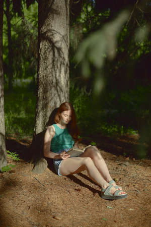 Redhead beautiful european woman sitting in the forest and reading a book.の写真素材