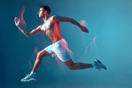 Running athletic male with naked torso in air on blue background in neon light. Motion blur. Modern...