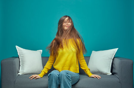 Modern young girl shaking her long brown tousled hair, enjoying haircare result, sitting on sofa. Natural hairs beautyの写真素材