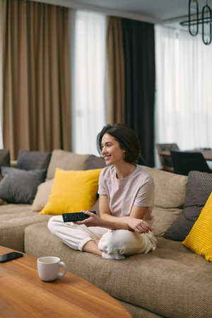 Happy young european girl watching television, enjoying TV program, series or movie, sitting on couch at homeの写真素材