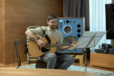 Talented young guy musician playing acoustic guitar during rehearsal, looking at sheet music sitting in musical studioの写真素材