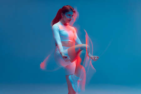 Isolated girl in white training suit on blue background. Leg swing with hands down. Fitness model in studio with long exposure effect.の写真素材