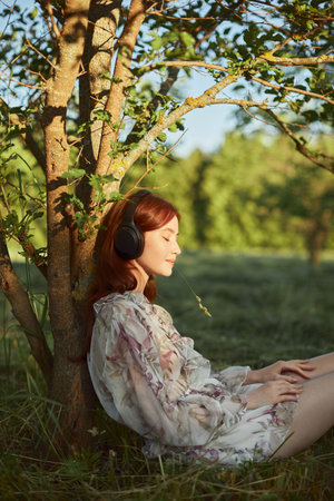 Young nice looking woman with ginger hair listening to the music with her eyes closed under the tree. Countryside landscape. Beautiful girl wears summer white dress.の写真素材