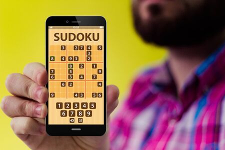digital gaming concept: Hipster with beard and checked shirt holding a smartphone with sudoku application form on the screenの写真素材