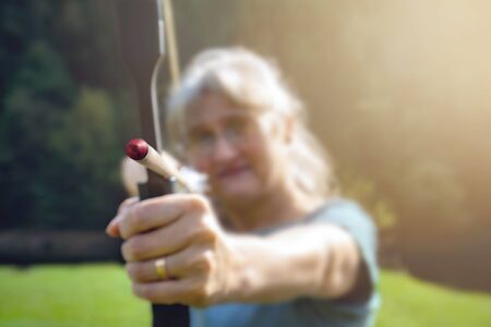 Woman with gray hair aiming with bow and arrow in nature and sun. Concept for the fight to reach the targetの写真素材