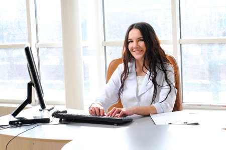 Portrait of young female doctor working in office, sitting at desk doing paperwork, looking at camera, smiling の写真素材