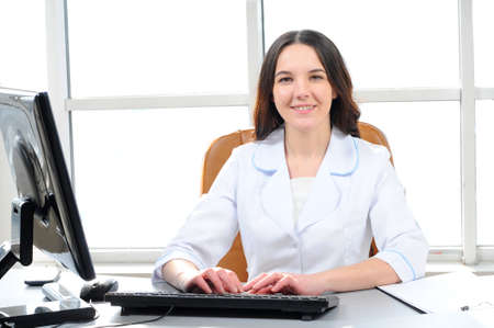 Portrait of young female doctor working in office, sitting at desk doing paperwork, looking at camera, smiling の写真素材