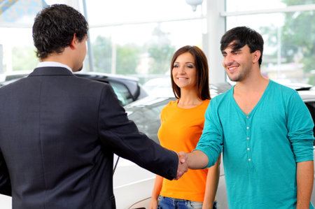 Sales situation in a car dealership, the dealer is talking to a young couple, there are cars standing in the backgroundの写真素材