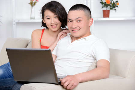 Portrait of young Asian couple sitting on sofa at home, using laptop computer, smiling.の写真素材