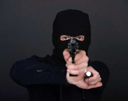 Masked robber with gun aiming into the camera against a black backgroundの写真素材