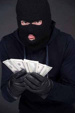Criminal in a balaclava holding a fistful of money conceptual of the loot from a robbery bribe corruption coercion payoff ransom or mob protection moneyの写真素材