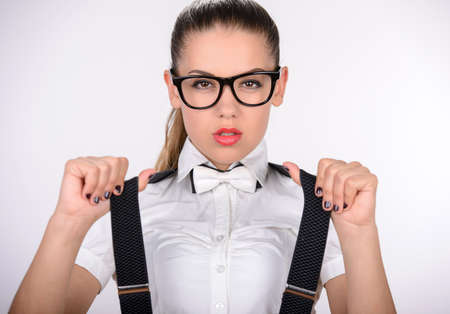 Always looking trendy. Beautiful young woman in glasses in white shirt and jeans shorts adjusting her suspenders and smilingの写真素材
