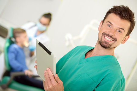A portrait of a dentist is looking at the camera with tablet, the dental assistant is working in the background.の写真素材