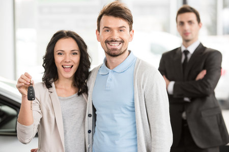 Happy young couple with keys from new car after automobile purchase in dealer showroom.