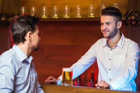 Cheerful young bartender is giving a mug with beer to the customer.の写真素材