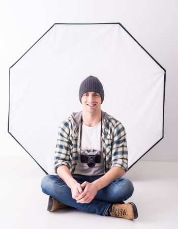 Young smiling photographer with camera in professionally equipped studio is sitting on the floor.