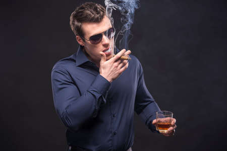 Young man in glasses is smoking a cigarette and is drinking whiskey over dark background.の写真素材