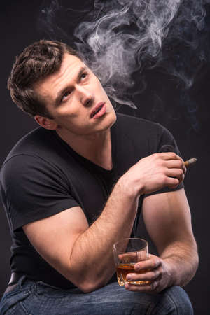 Young man is smoking a cigarette and is drinking whiskey over dark background.の写真素材