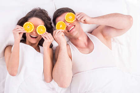 Top view. Young smiling couple lying on bed nad putting on eyes slices orange.の写真素材