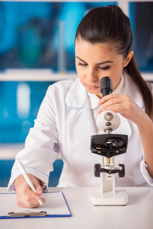 Attractive female scientist analysing something through a microscope and doing notes in a...
