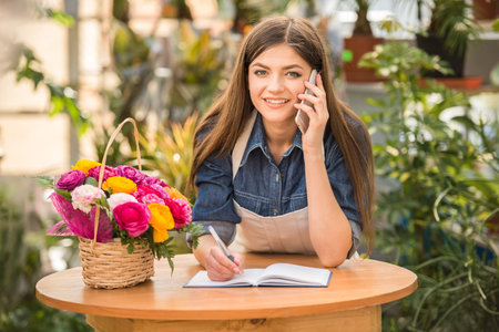 Portrait of young female florist talking on phone and making notes at flower shop.の写真素材