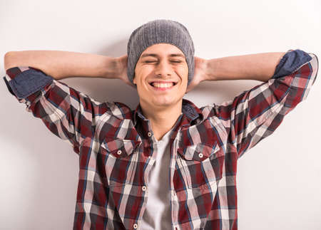 Handsome young man is holding hands behind head and smiling while standing against gray background.の写真素材