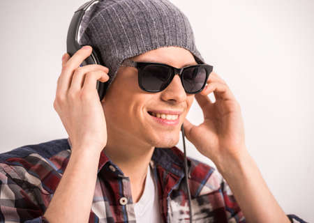 Happy young stylish man in sunglasses and headphones is smiling while standing against grey background.の写真素材