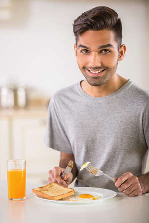Young handsome man in grey t-shirt eating omelet and drinking juice for breakfast.の写真素材