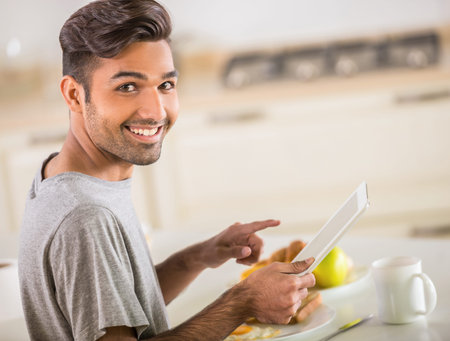 Young handsome man in grey t-shirt eating breakfast and using digital tablet.の写真素材