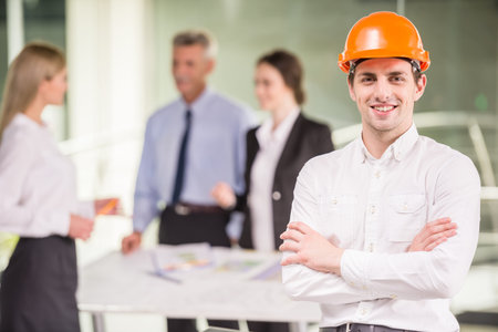 Happy architect in orange helmet standing at office with team on background.