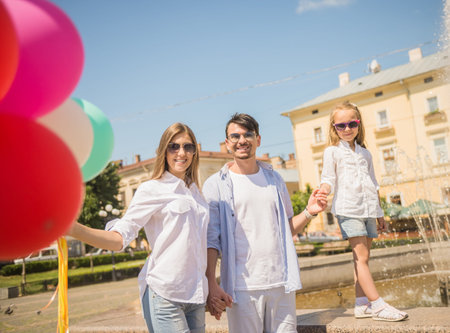 Happy young family walking along the street of european city with  bunch of colorful balloons, fountain on background.の写真素材