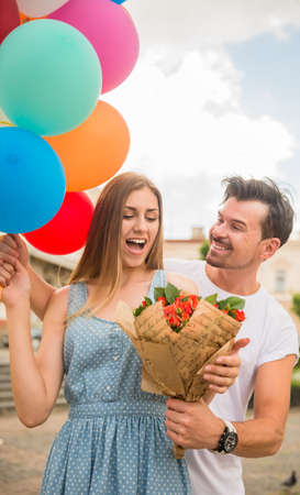 Romantic date outdoors. Handsome man surprised his woman with flowers and colorful balloons. Front view.の写真素材